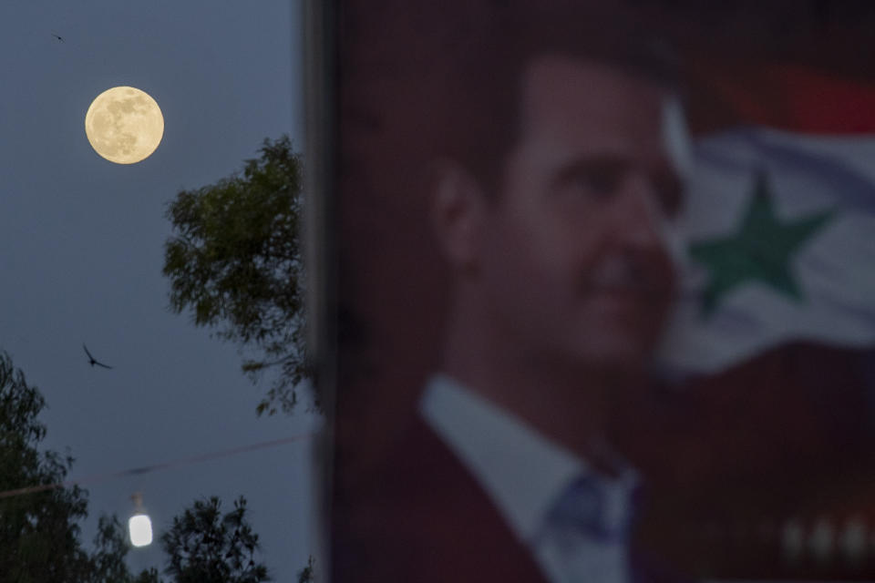 A full moon rises behind a campaign poster of President Bashar Assad for the upcoming presidential election that adorn a street in the Syrian capital Damascus , Syria, Tuesday, May 25, 2021. The presidential elections in the war-ravaged country will be held May 26. (AP Photo/Hassan Ammar)