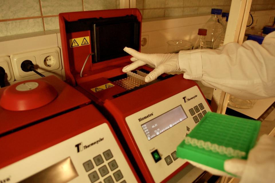 Una persona en ropa quirúrgica blanca con guantes blancos coloca muestras dentro de un dispositivo de laboratorio rojo.