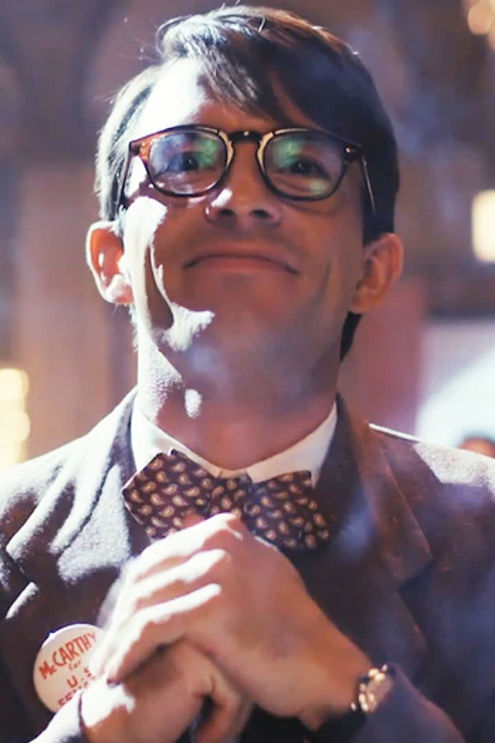 Jonathan Bailey wearing glasses, a bow tie, and a dark suit smiles and clasps his hands together. A crowd and chandelier are visible in the blurred background