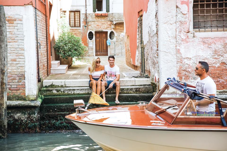 Young couple sit on stone steps next to river while speedboat passes in Venice, Italy