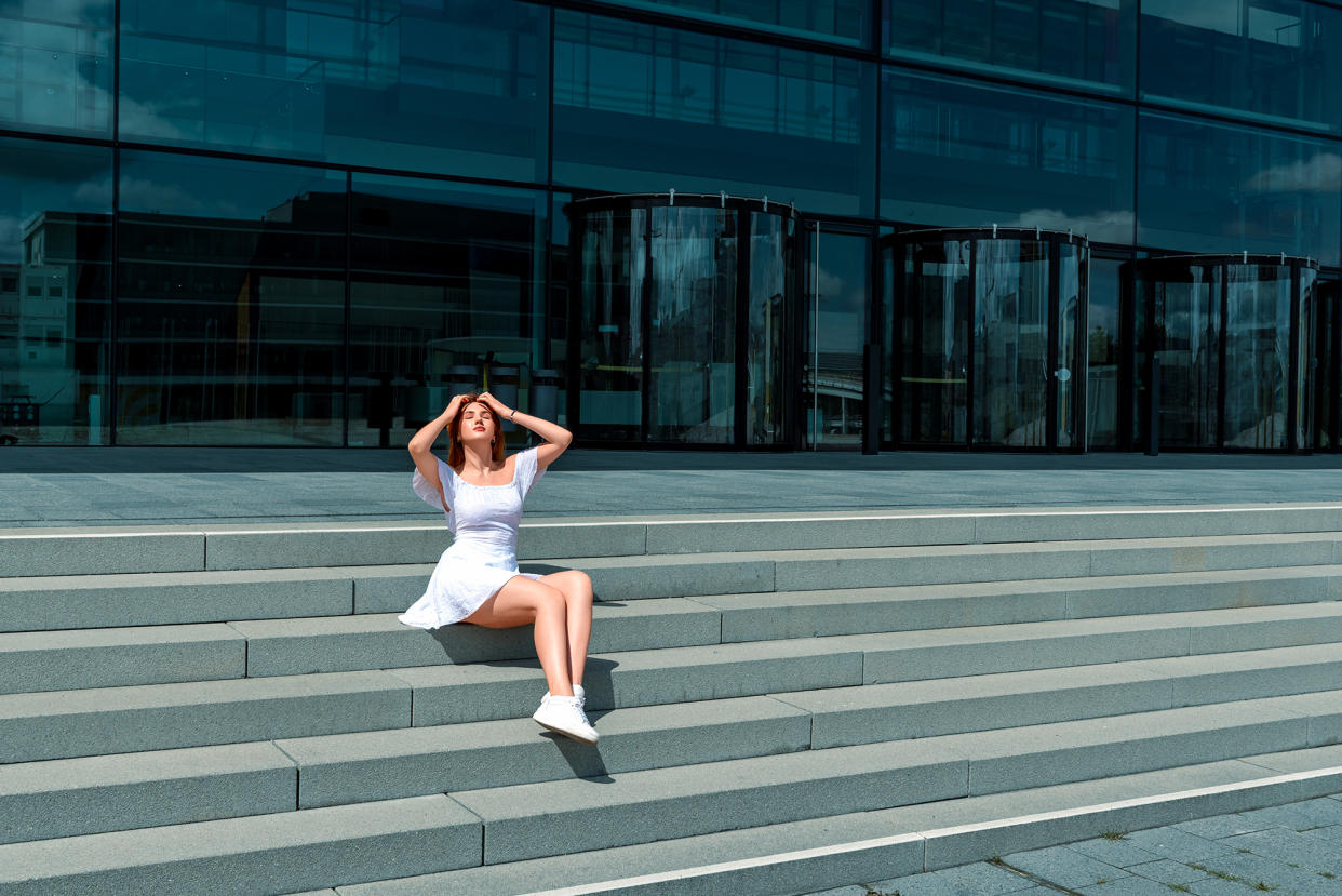 Beautiful young woman sitting on stairs and enjoys the bright sunlight on a summer day. Young woman wearing a white dress and sneakers sitting on stairs in front of a hotel