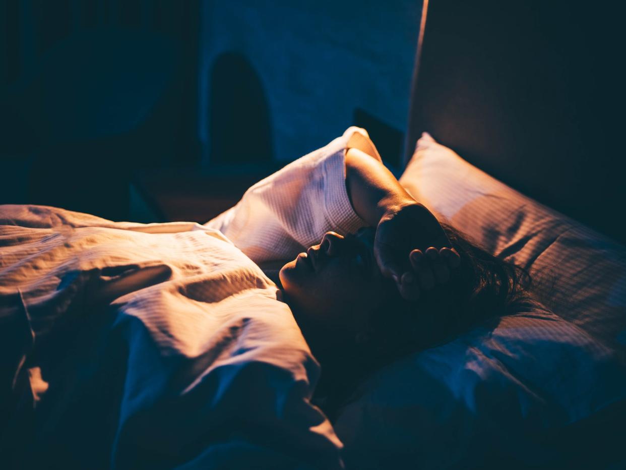 Photo from the side of a woman lying in bed, staring at the ceiling with her right arm across her head and the blanket pulled up to her chin. The woman has dark hair and is lit by a warm overhead lamp and a blue light from outside. 