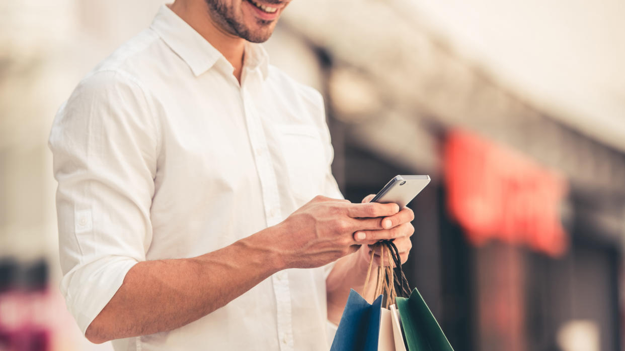 Guy with shopping bags is using a mobile phone and smiling while doing shopping in the mall.