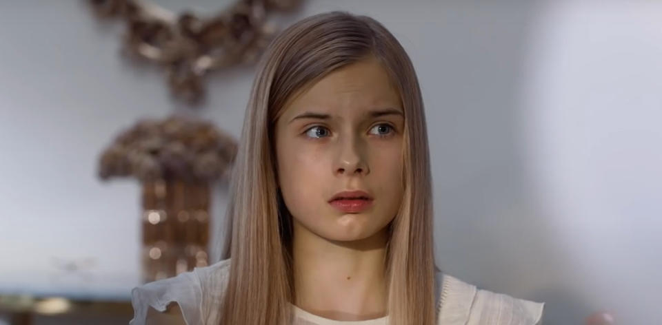 Tinja, played by Siiri Solalinna, looks slightly to the side with a concerned expression. A blurred, ornate mirror and vase are in the background