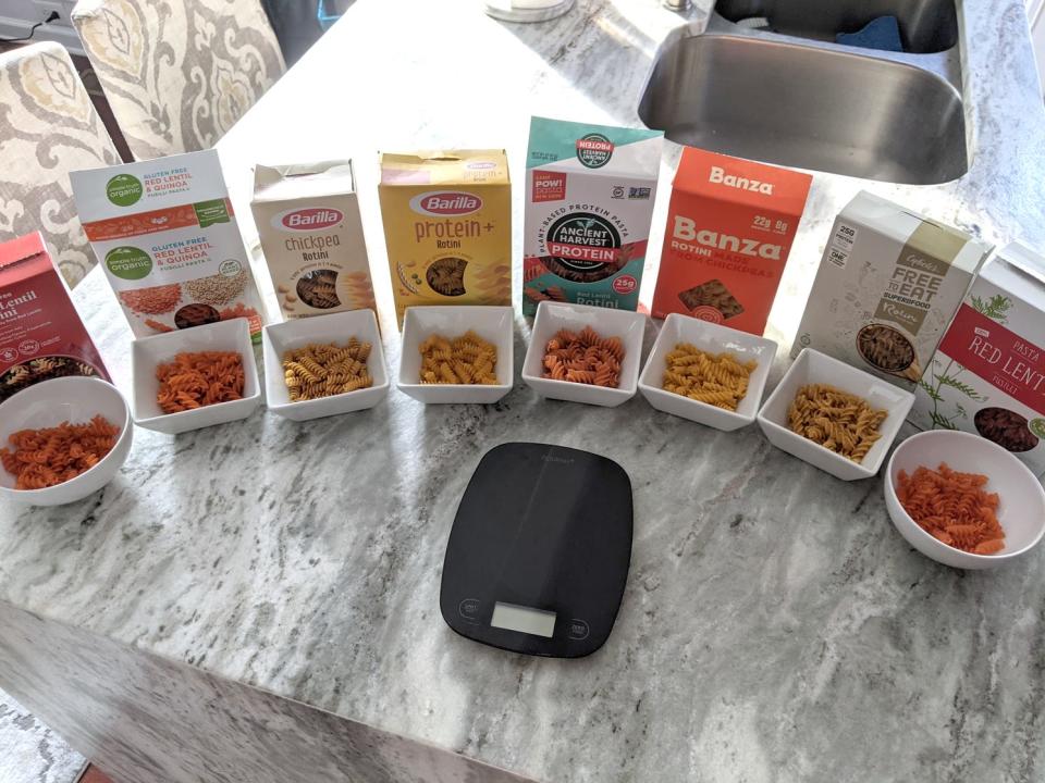 eight boxes of different brands of protein pasta lined up on a kitchen counter next to a kitchen scale