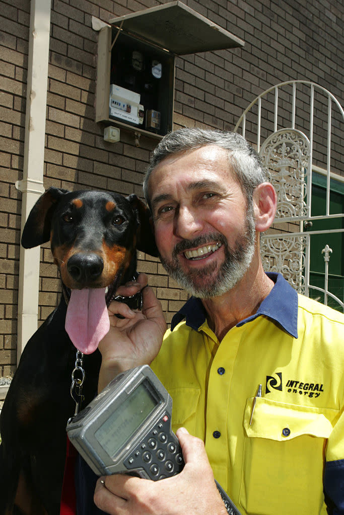 A man in a yellow and blue Integral Energy shirt smiles while holding a device. A black Doberman with its tongue out is next to him