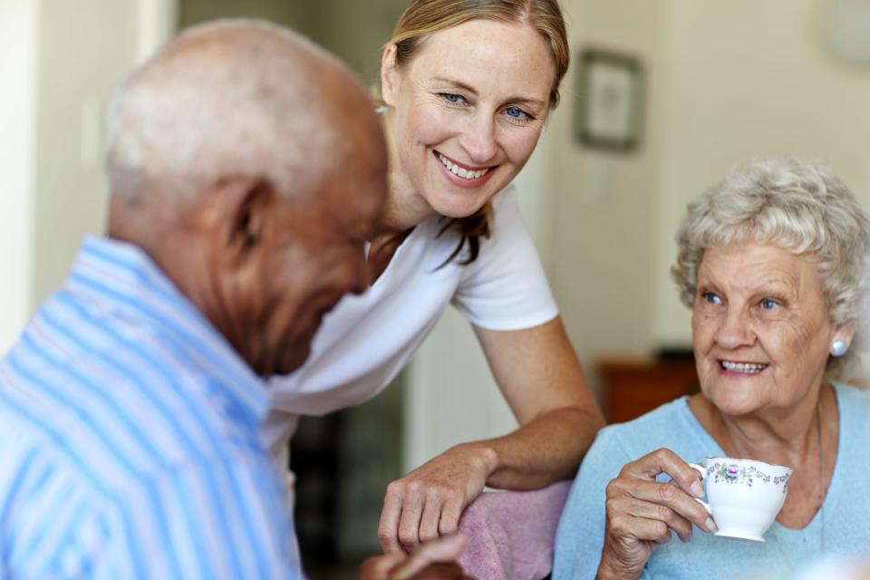 With a smiling staff member looking on, two nursing home residents enjoy conversation while having coffee.