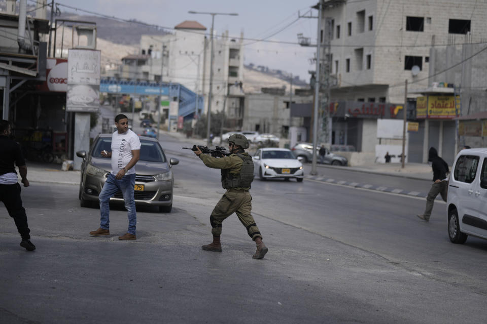 An Israeli soldier draws his weapon on Palestinians, left, as settlers, right, attack shops in Huwara, near the West Bank town of Nablus, Thursday, Oct. 13, 2022. (AP Photo/Majdi Mohammed)