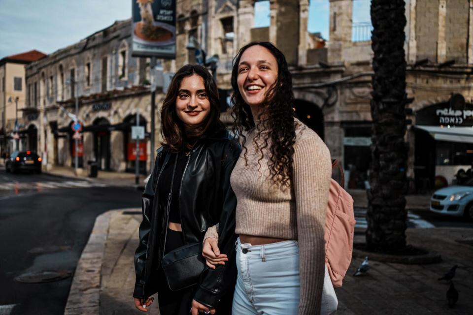 Two teen girls stand close together smiling on a street