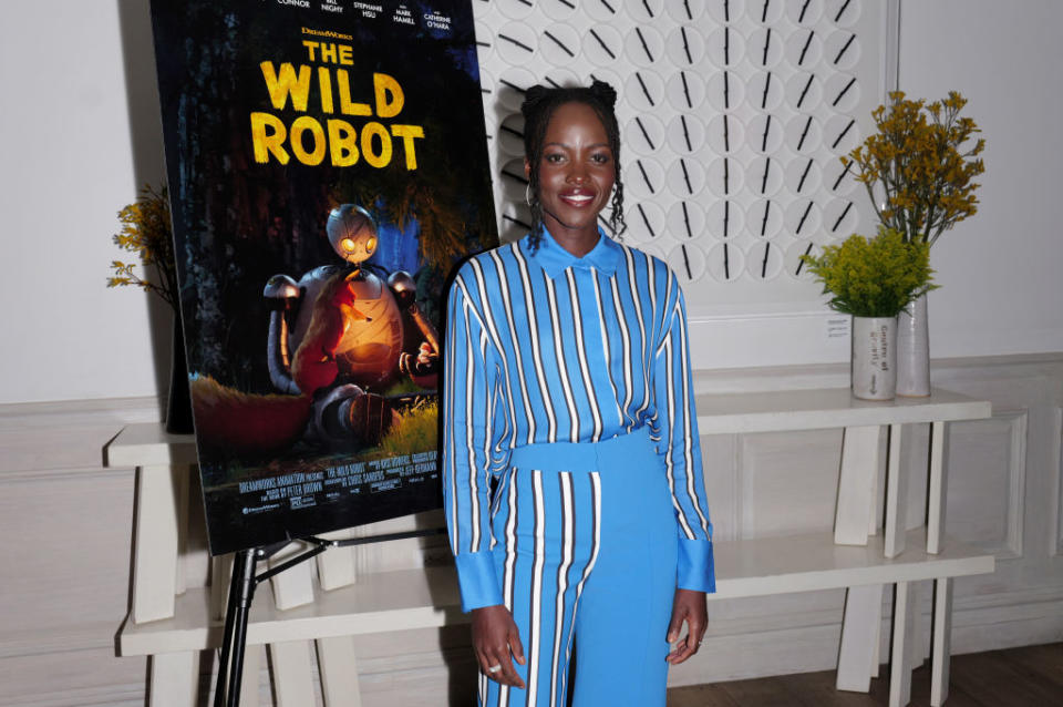 Lupita Nyong'o poses in front of a poster for the film "The Wild Robot," wearing a stylish striped jumpsuit with braids