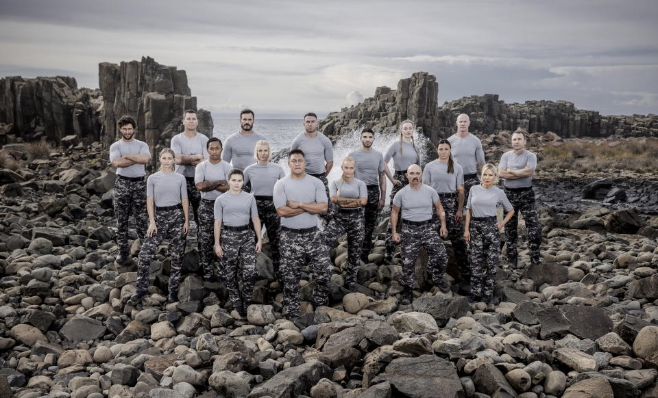 Group shot of 17 SAS Australia celebrity recruits at Bombo Quarry in the Illawarra region of NSW, Australia. Men and women in camouflage pants and grey t-shirts stand in three rows with the sea behind them on rocks. 