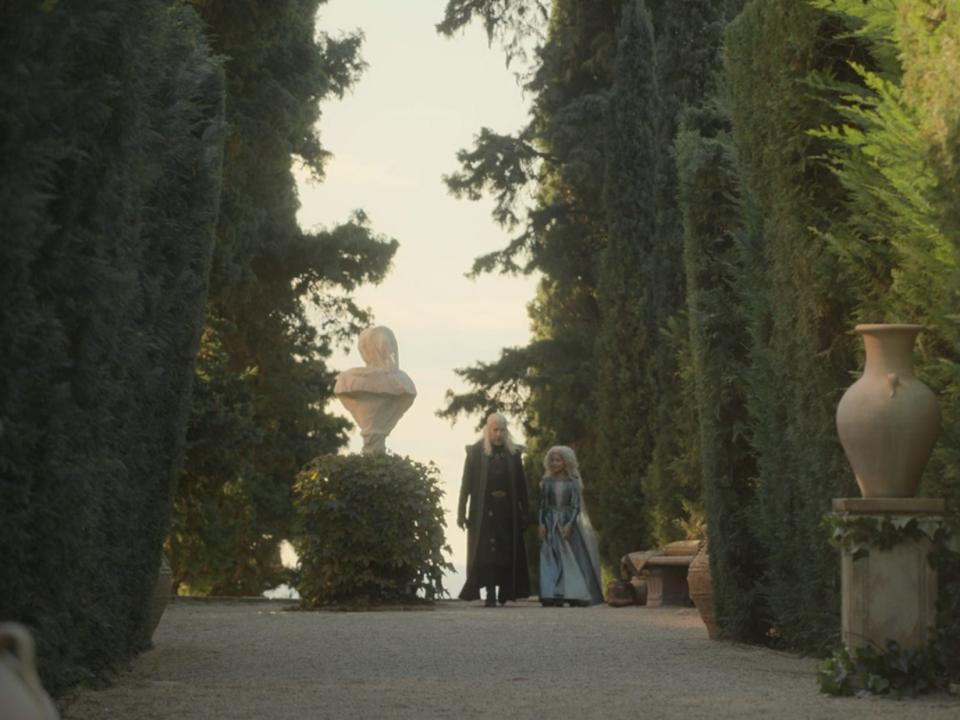 A scene from "House of the Dragon," showing a man and a young girl walking together in a garden with high hedges lining the pathway.