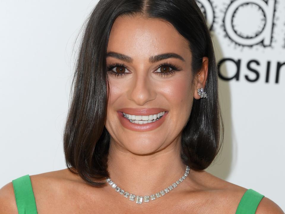 Lea Michele, in a green strapless dress and diamond jewelry, smiles while posing on the red carpet.