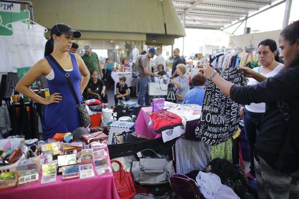 In this Nov. 3, 2019 photo, Yenika Calderon, left, watches customers look through her personal belongings at a secondhand market, in Caracas, Venezuela. Calderon, 41, who is hoping to pocket enough money to give her young family a fresh start far away in Spain, has trekked to the secondhand market every Sunday in recent weeks, haggling with customers over prices for her favorite handbags and her son’s baby clothes. (AP Photo/Ariana Cubillos)