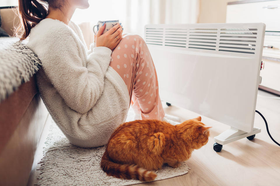 Woman warming and drinking tea with cat sitting by device and wearing warm clothes. 