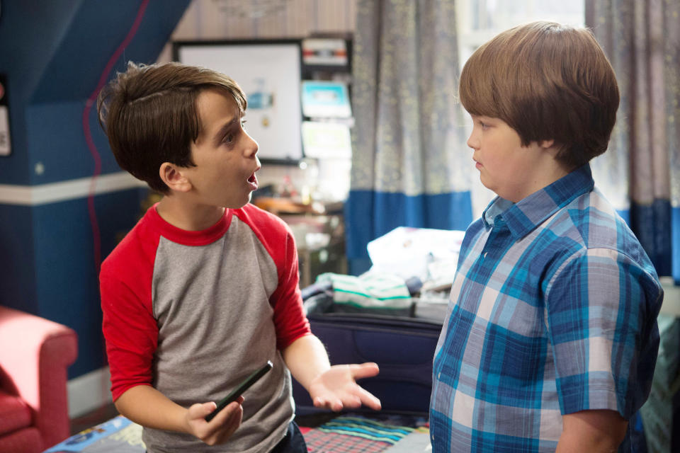 Zachary Gordon and Robert Capron in a room talking, one gestures while holding a smartphone, the other listens with hands in pockets