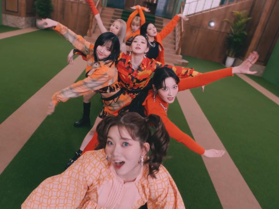 the seven members of billlie, young women dressed in orange outfits, dancing. they're arranged in a line between one woman in the front, spread out to the sides to show each members' face and extending their arms outward
