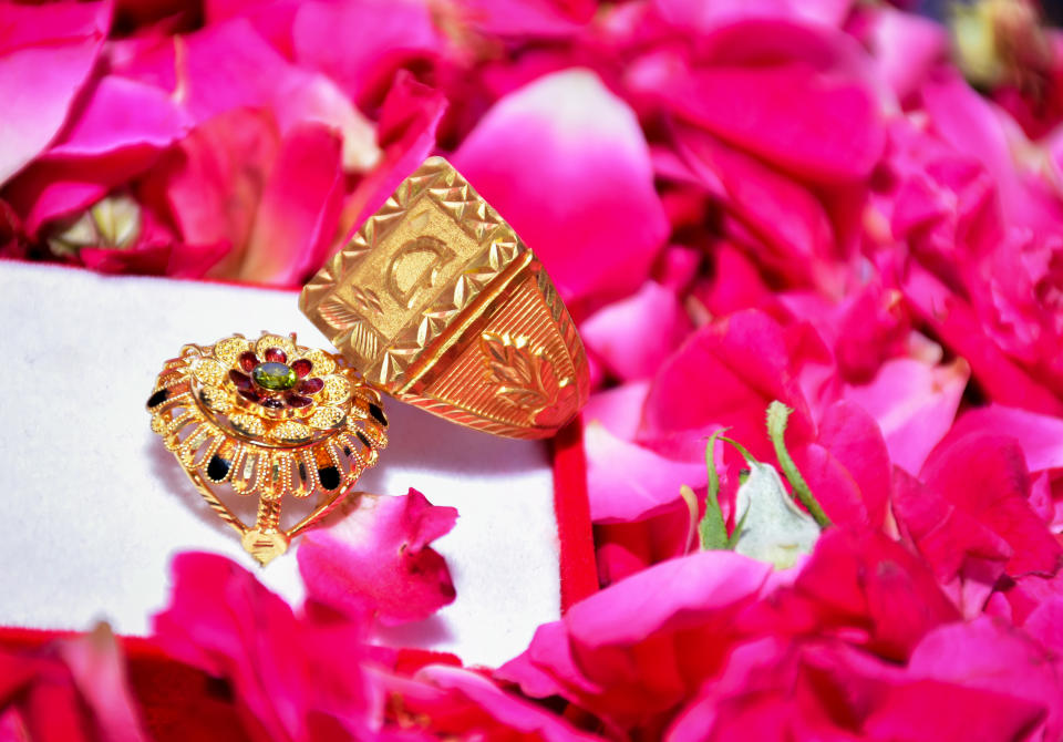 Two intricately designed gold rings rest on a bed of rose petals at Roka. One ring features a large rectangular design, while the other is ornate with red and black gemstones