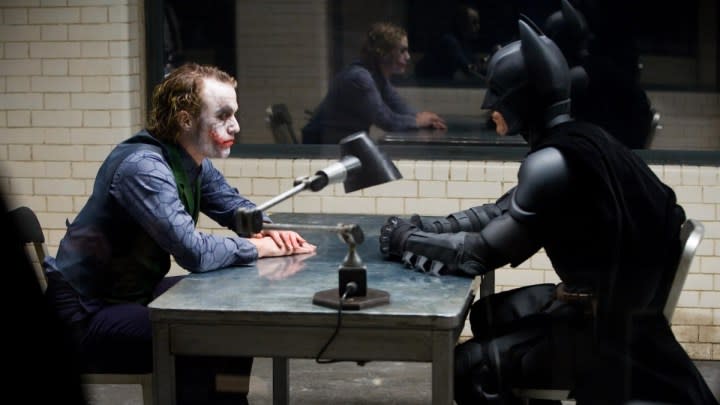 The Joker and Batman sit across from one another at a table.