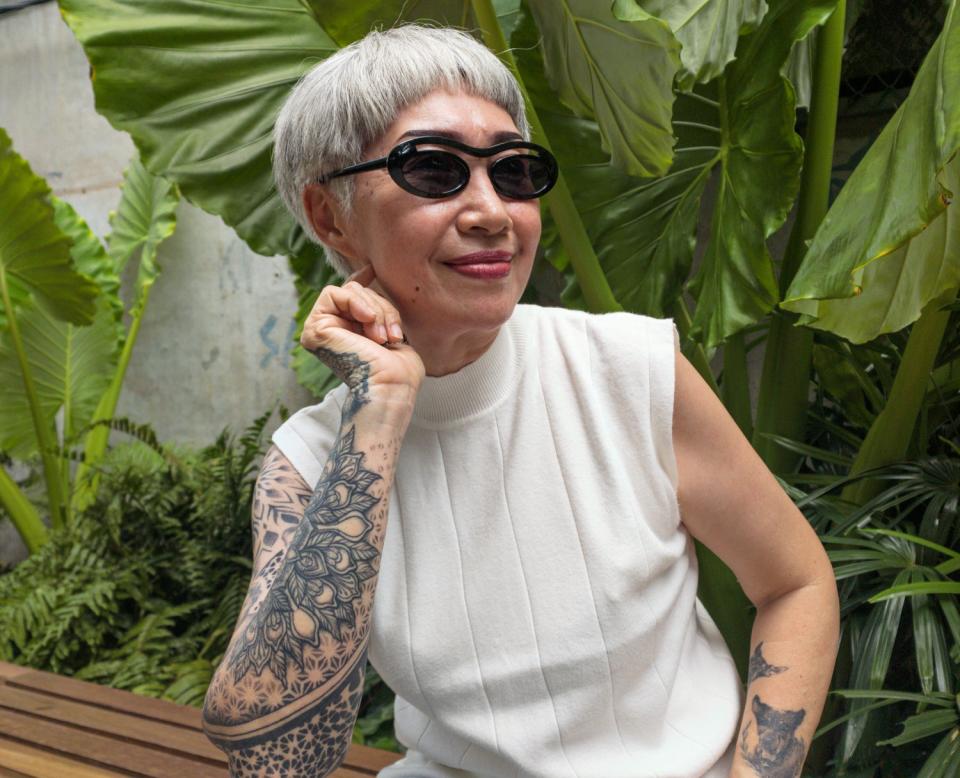 An elderly woman with tattoos smiles while sitting on a bench, wearing sunglasses, a sleeveless white top, and jeans, in a lush garden setting