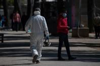 A Health Ministry official wearing a protective suit walks past a migrant wearing a protective face mask and standing in the Ritsona camp, after authorities found 20 coronavirus cases and placed the camp under quarantine, in Ritsona