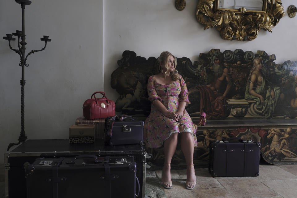 a woman (jennifer coolidge as tanya mcquoid) sits at a bench painted with artwork, next to a black trunk and several pieces of luggage