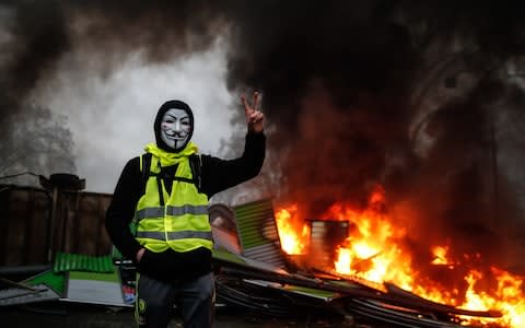 A protester wearing a Guy Fawkes mask makes the victory sign near a burning barricade in Paris during a protest of yellow vests (gilets jaunes) against rising oil prices and living costs - Credit: Abdulmonam Eassa/AFP