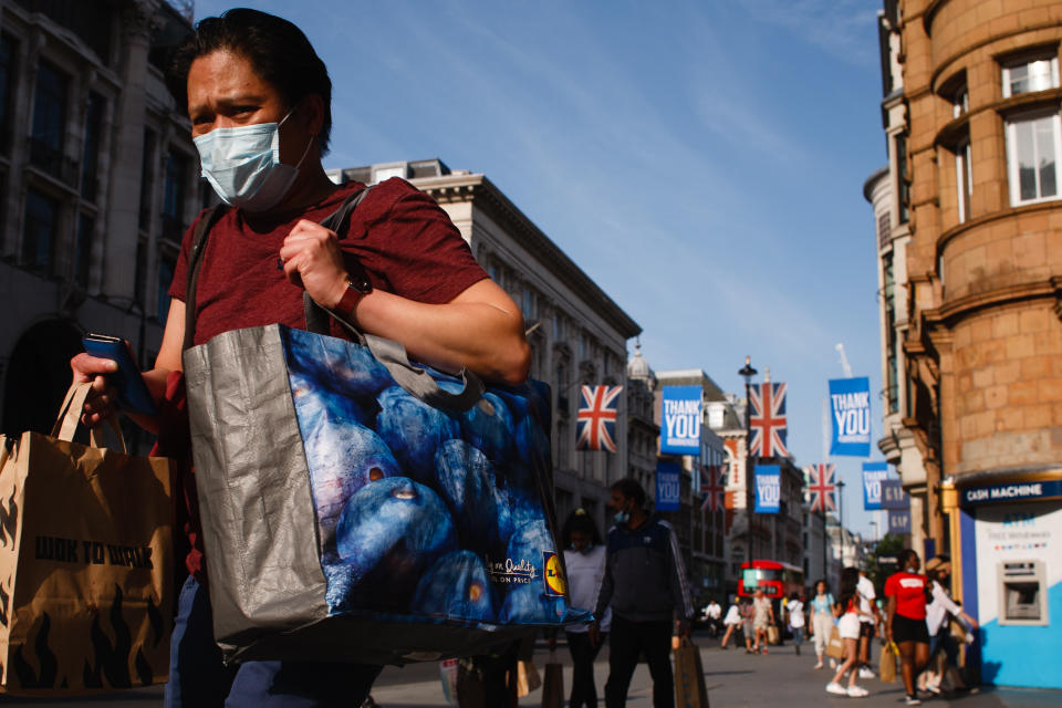 A shopper wearing a face mask carries bags along Oxford Street in London, England, on June 23, 2020. British Prime Minister Boris Johnson announced today that the next stage of lockdown easing in England would proceed on schedule, with pubs, restaurants, hotels, hairdressers, theatres, cinemas, museums, galleries, libraries, theme parks and zoos allowed to reopen from July 4. The two-metre social distancing rule is also to be halved from the same date, with people encouraged to take additional mitigation actions, such as wearing face coverings, when close together. The change, to what is being dubbed 'one-metre plus', is seen as key to the survival of the hospitality sector. (Photo by David Cliff/NurPhoto via Getty Images)