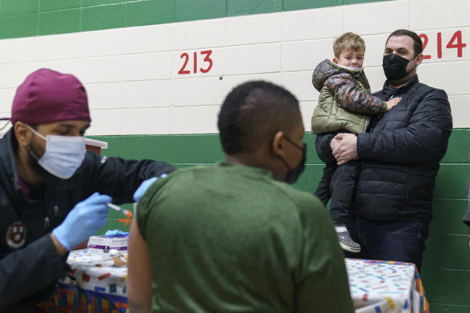 Brian Stachowiak, right, holds his son, Noah, 5, as he watches another child receive a COVID-19 vaccination from Dr. Eugenio Fernandez, while waiting his turn at Nathanael Greene Elementary School in Pawtucket, R.I., Tuesday, Dec. 7, 2021. Even as the U.S. reaches a COVID-19 milestone of roughly 200 million fully-vaccinated people, infections and hospitalizations are spiking, including in highly-vaccinated pockets of the country like New England. (AP Photo/David Goldman)