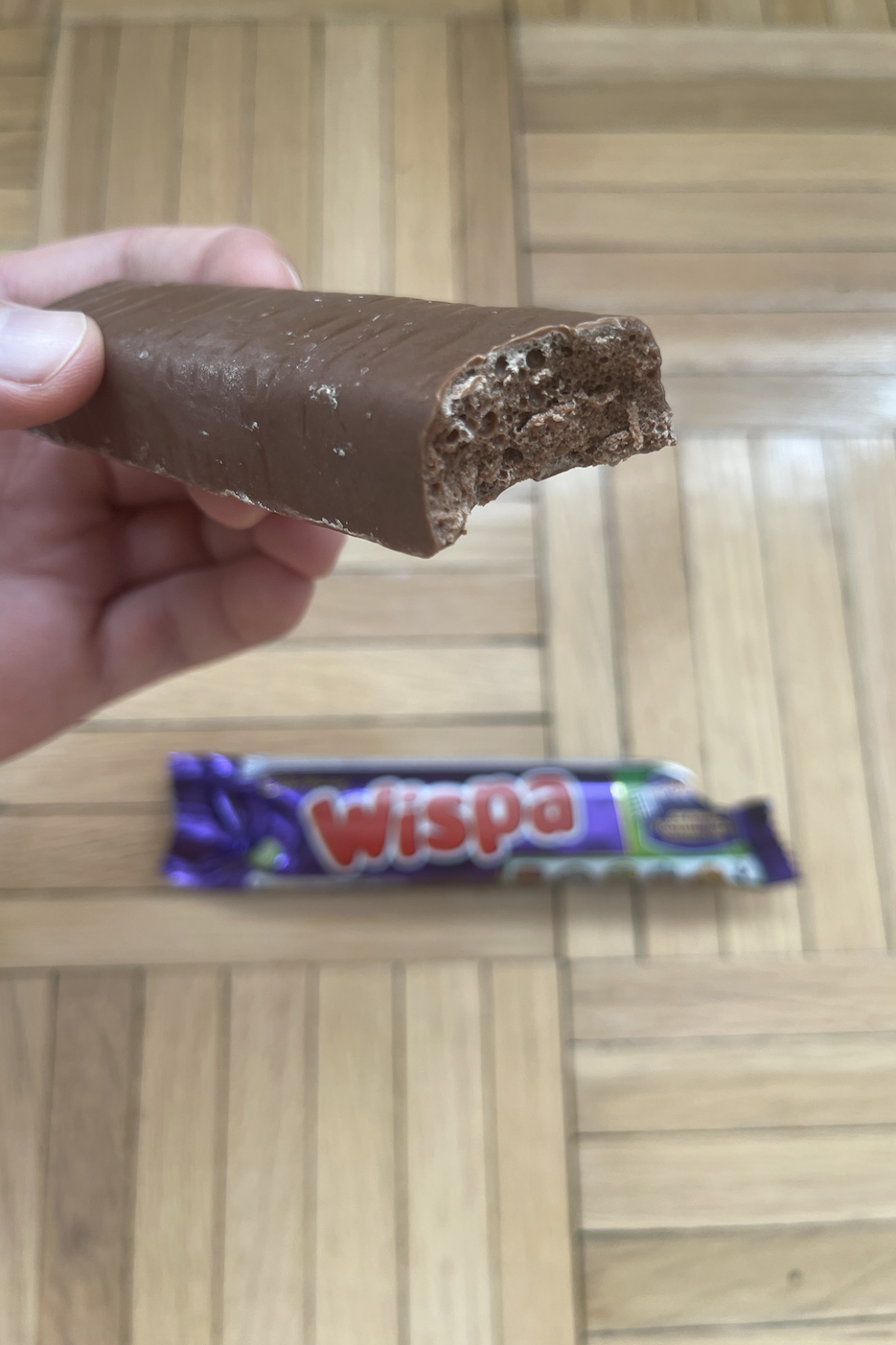 Person holding a half-eaten Wispa chocolate bar with another unopened bar on a wooden floor