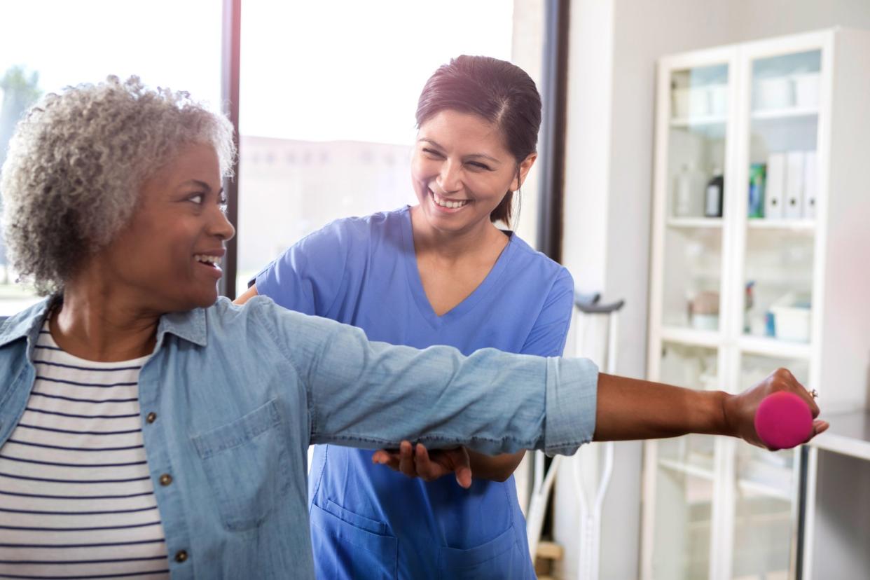 A smiling senior woman sits in a physical therapy office.  She reaches out to exercise her arm with a hand weight as her cheerful mid adult female physical therapist stands behind her and guides her arm.