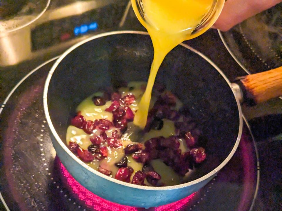 Cranberries in a saucepan with a hand pouring orange juice into the pot above the fruit. The pan sits on an electric burner.