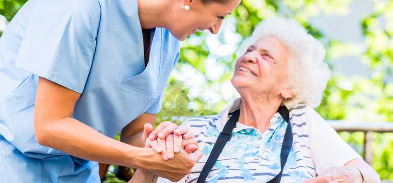 A health care worker holds hands with an elderly woman in wheelchair.