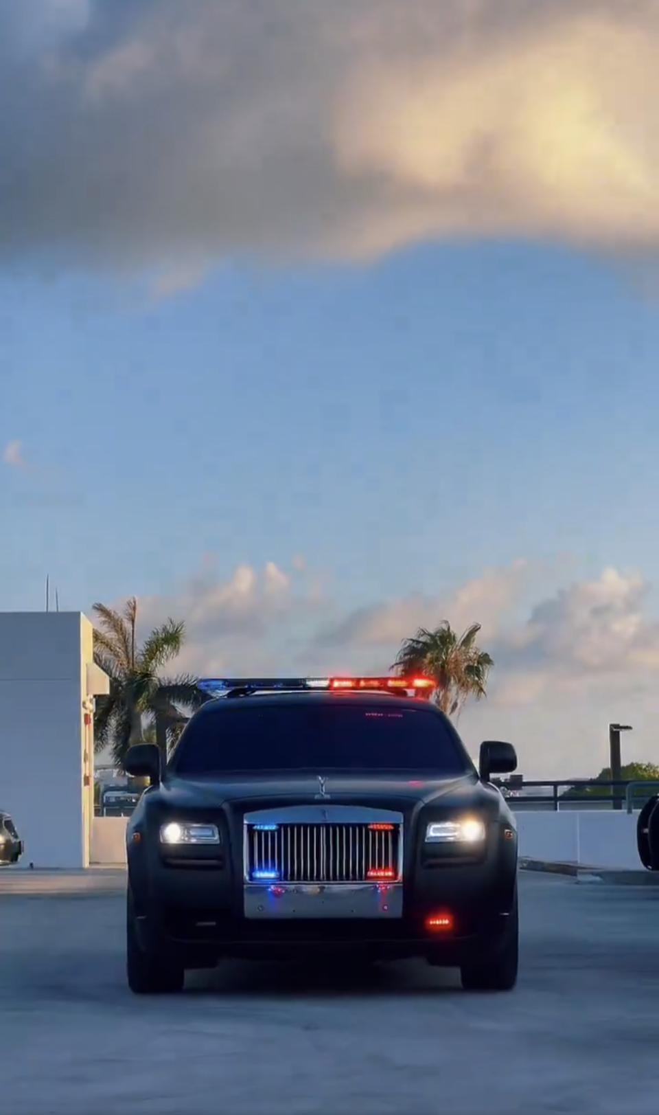 A luxury car with police lights is parked near a building with palm trees in the background, capturing a newsworthy moment