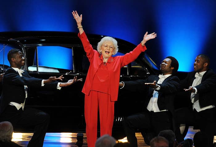 Betty White with her arms in the air onstage as she's surrounded by background dancers in tuxedos