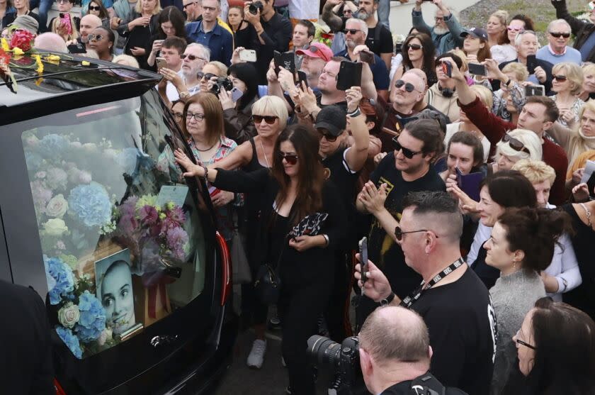 A crowd of people in black surround a hearse filled with pink and blue flowers and a picture of late singer Sinéad O'Connor