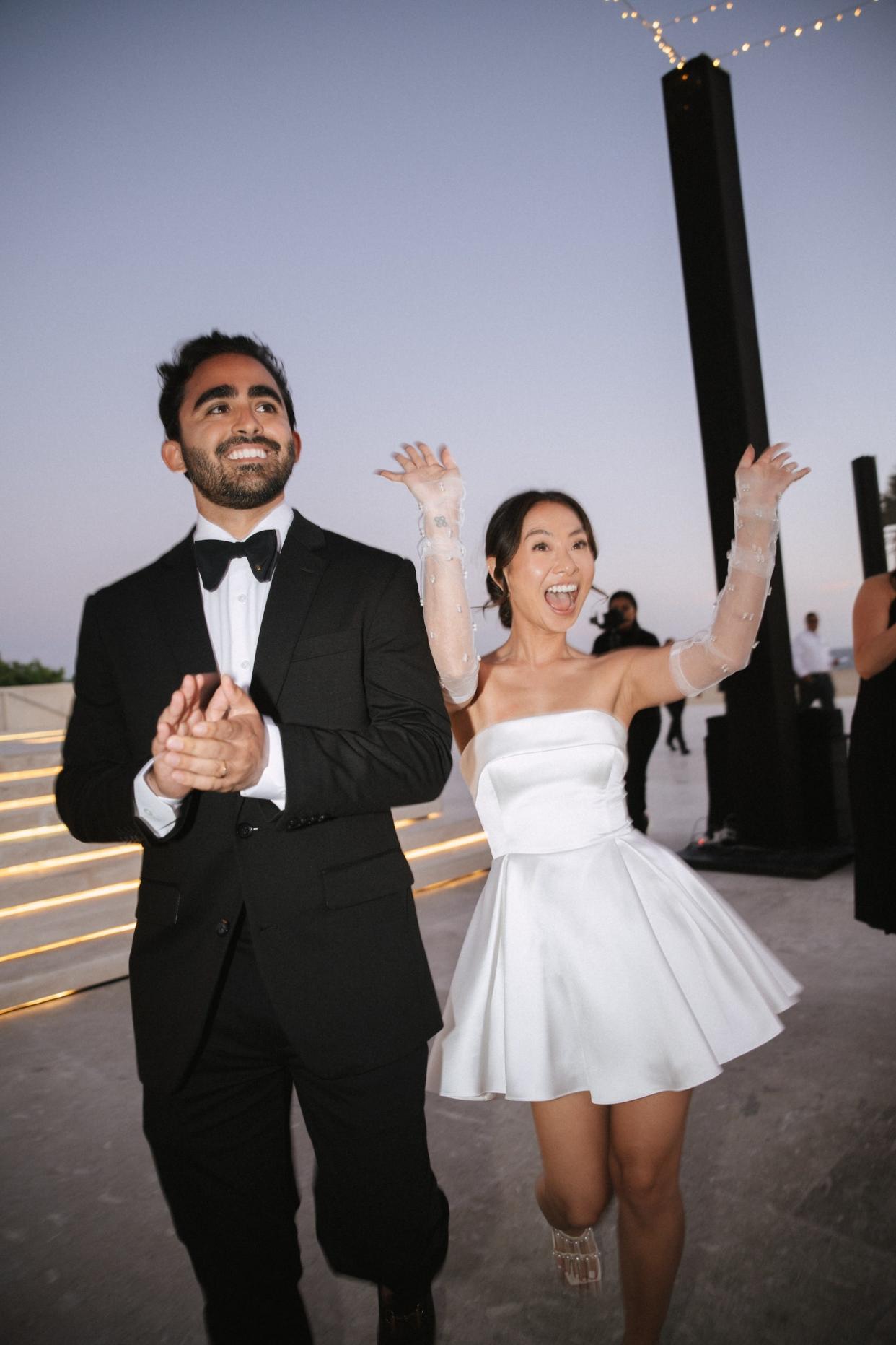 A bride and groom walk into their wedding reception. The bride waves in a shorter dress.
