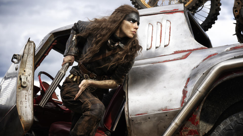 Furiosa loads a gun as she steps out of a moving vehicle's side door in Furiosa: A Mad Max Saga