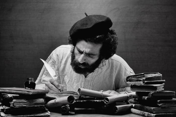 A man with a thick beard and a beret intensely writes with a quill pen at a cluttered desk filled with books, scrolls, and an inkpot