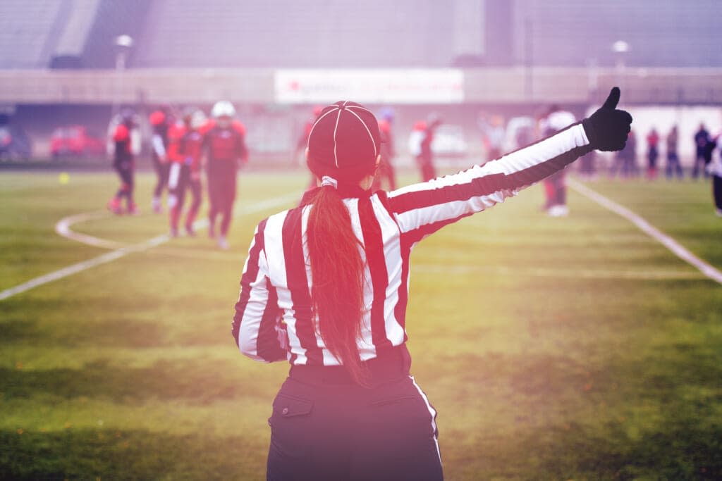 Female American football referee giving signals to professional players during match on the stadium field. (Adobe Stock)