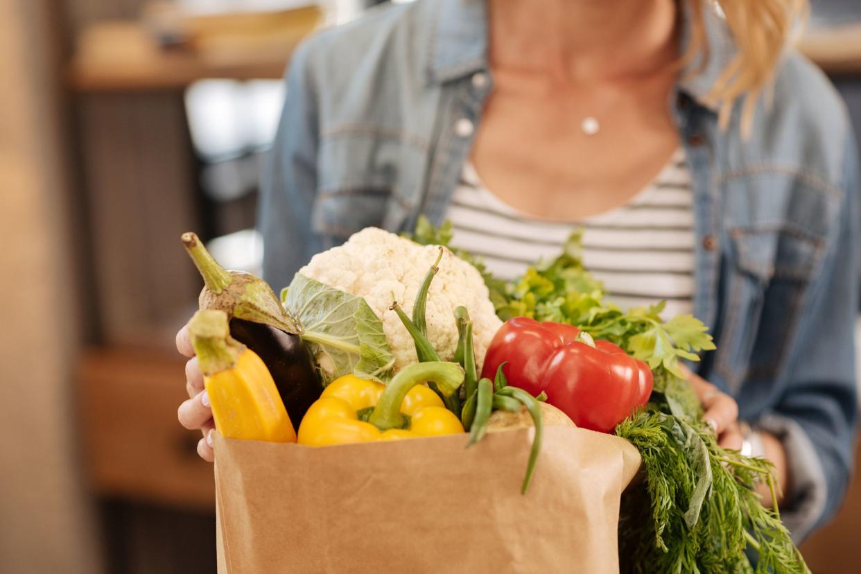 attractive and fit middle-aged woman unpacking brown bag of grocery items, mostly fresh vegetables, on the table of her kitchen