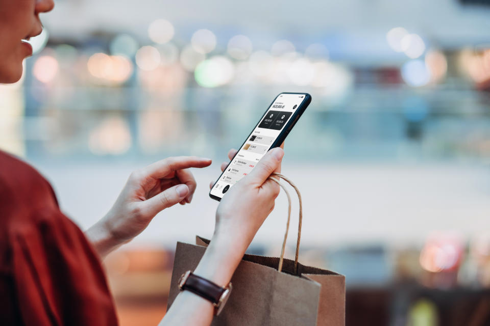 Cropped shot of young Asian woman holding a paper shopping bag, managing online banking with mobile app on smartphone. Transferring money, paying bills, checking balance while shopping in a shopping mall. Technology makes life so much easier