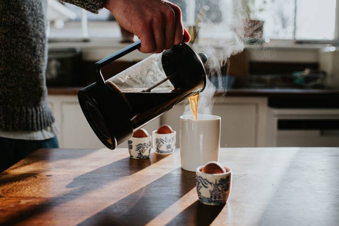 A person pours coffee from a French press into a mug on a kitchen counter. Three soft-boiled eggs in decorative cups are also on the counter