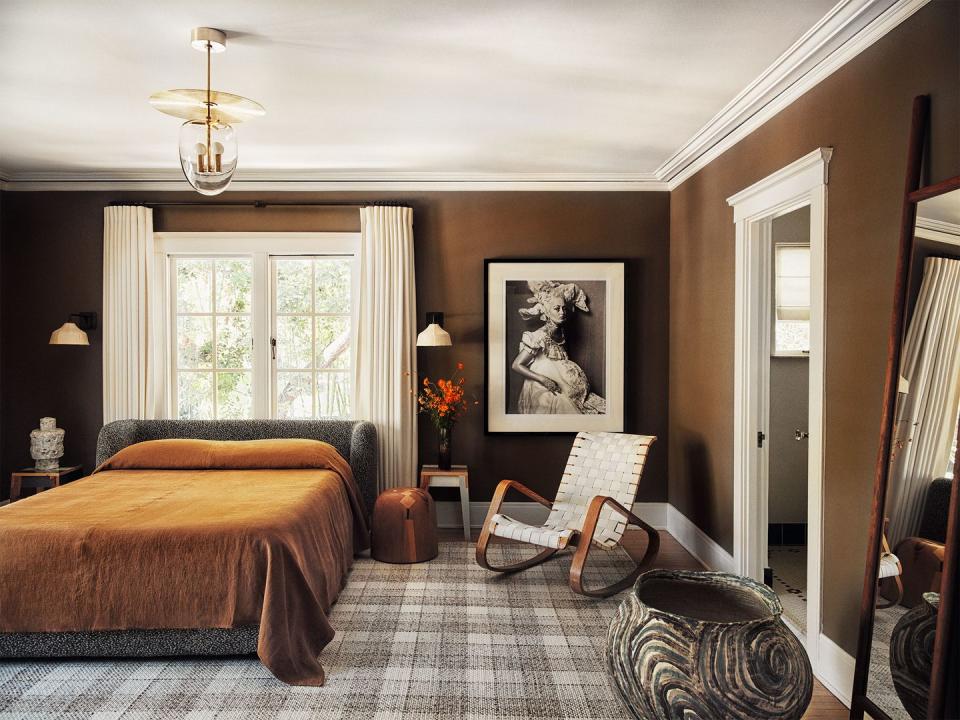 primary bedroom with medium chocolate painted walls and a plaid rug in neutral tones and a slatted rocking chair next to the bed with complementary coverlet in brown on the bed