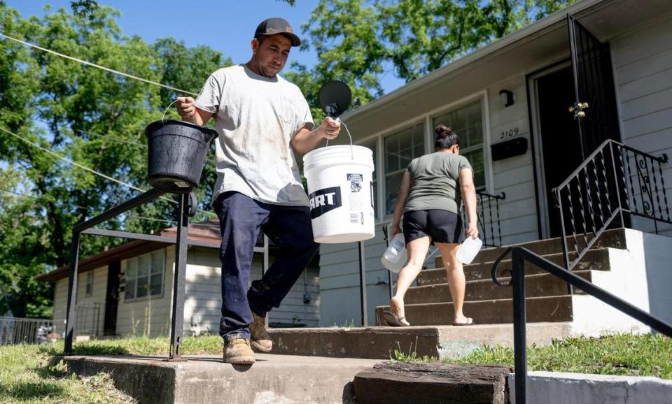 Alfonso López camina con cubetas vacías para traer agua de su auto mientras su esposa Jessica Martínez lleva jarras de agua al interior, el viernes 26 de mayo de 2023, en Kansas City. Debido a un saldo pendiente dejado por un inquilino anterior, KC Water cortó el agua a la casa de la pareja en abril. Desde entonces, su familia ha tenido que ser creativa para cubrir sus necesidades de agua, incluyendo el transporte en auto de viejas botellas de refresco y cubetas llenas de agua hasta su casa, y el uso de agua embotellada para cepillarse los dientes.