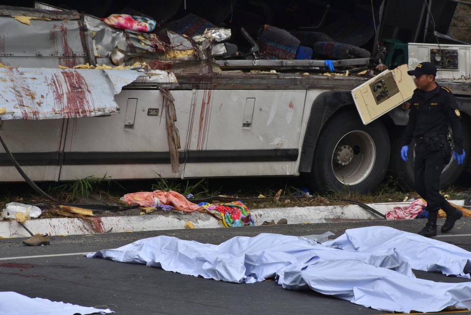 A police officer walks past the covered bodies of passengers that perished when a bus crashed with a trailer truck in Gualan, Guatemala, Saturday, Dec. 21, 2019. The accident killed at least 21 people and left a dozen wounded, according to the national disaster agency. (AP Photo/Carlos Cruz)