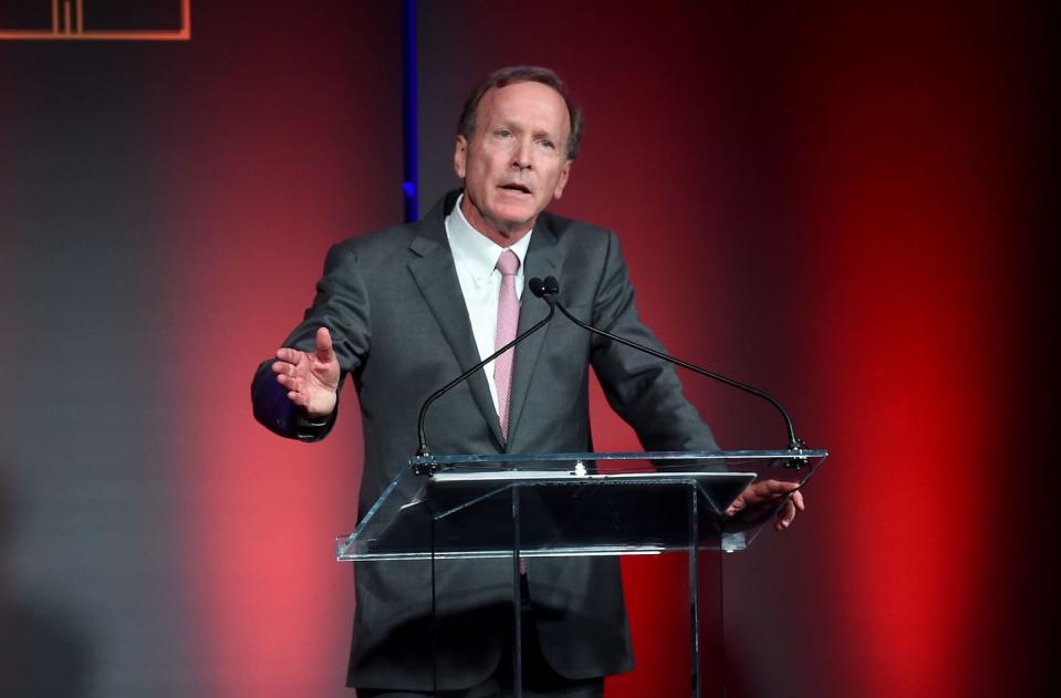 neil bush wearing a black suit and tie, standing at a podium and speaking into two microphones, in front of a red backdrop