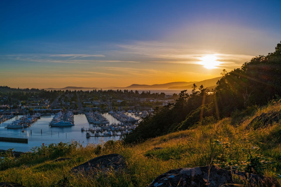 Sunset over a harbor filled with boats, viewed from a grassy hillside. The sky has a gradient from bright to dark as the sun descends behind distant mountains