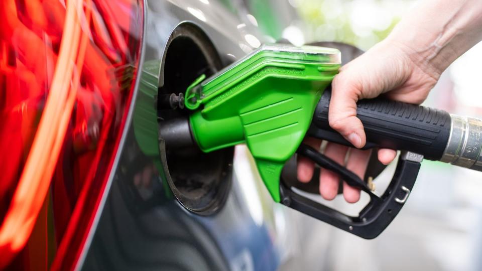 Eine Frau hält an einer Tankstelle an einer Zapfsäule eine Zapfpistole in der Hand und betankt ein Auto.