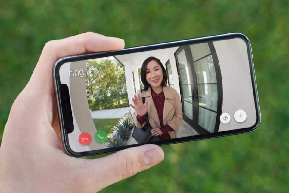 A close up of the screen of a smart phone that shows the security video footage of a woman waving at a video doorbell. 